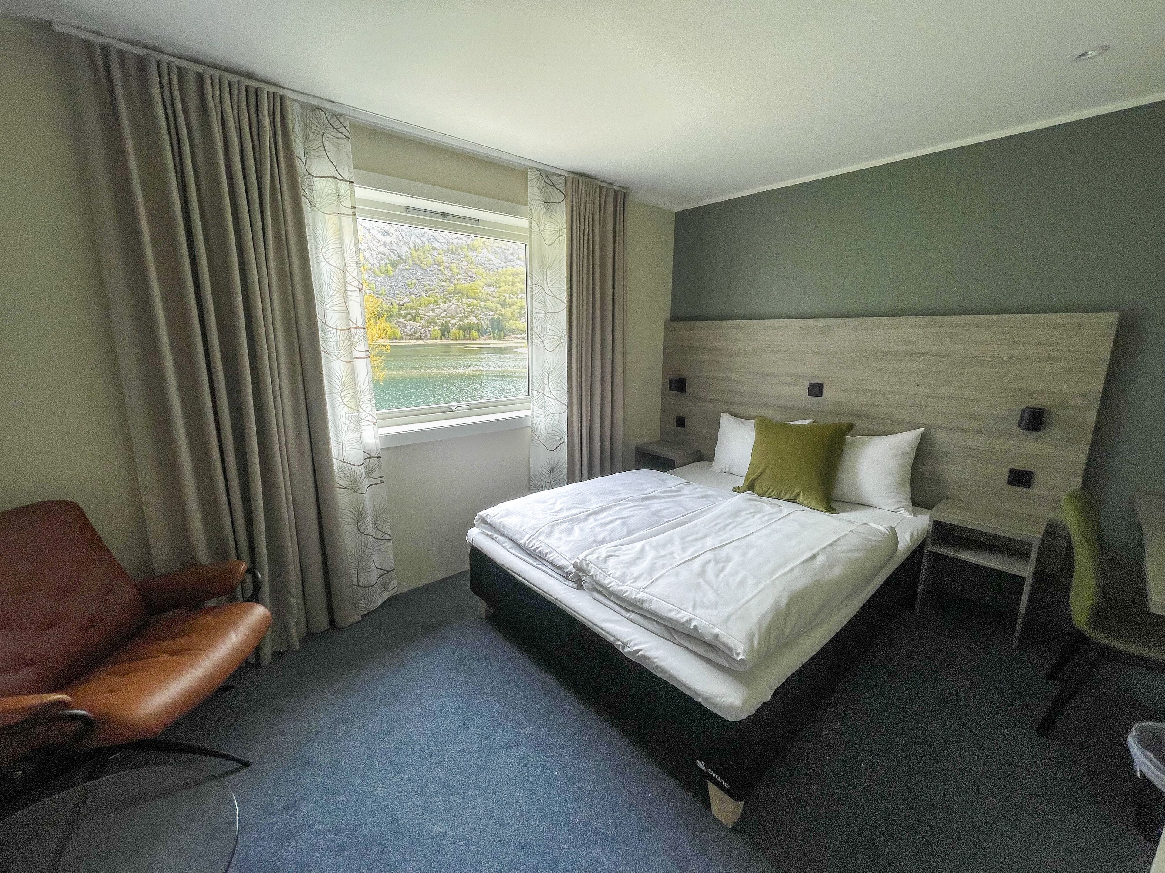 Une chambre d’hôtel avec un grand lit, une tête de lit en bois, une fenêtre avec des rideaux, un fauteuil en cuir marron et une petite table de chevet.