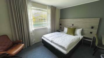 Habitación de hotel con una cama grande, cabecera de madera, ventana con cortinas, un sillón de cuero marrón y una mesita de noche pequeña.