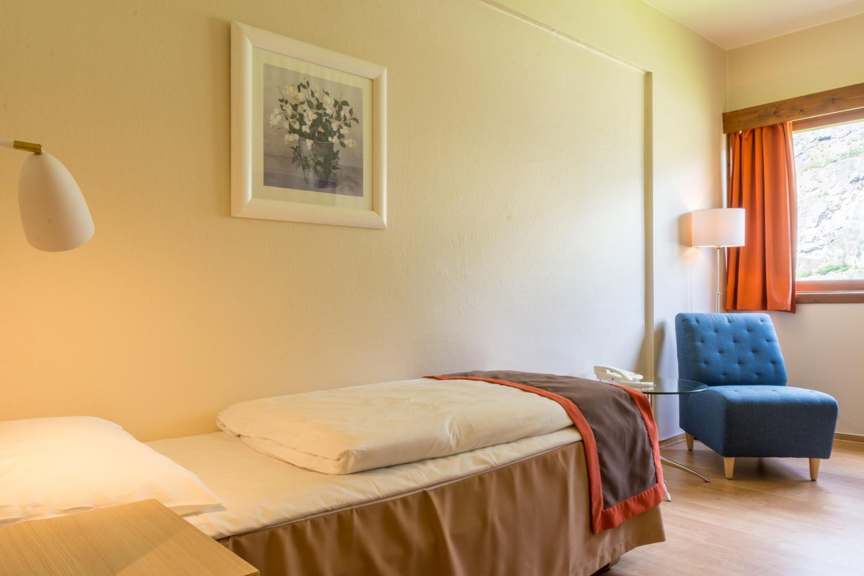 Une chambre d’hôtel avec un lit, une chaise, une table de chevet, une lampe et une fenêtre avec des rideaux.