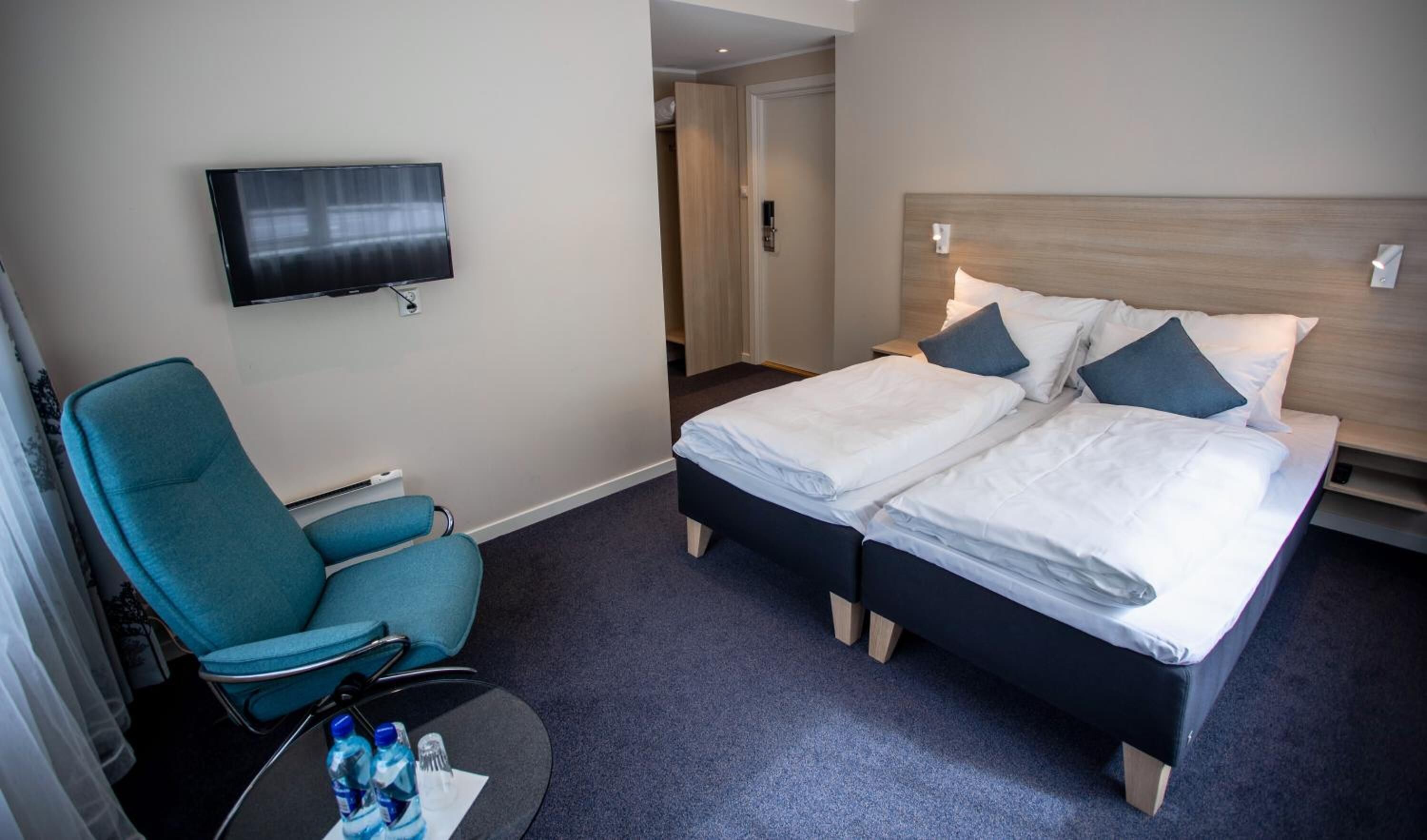 Et hotelværelse med seng, fjernsyn, stol og et lille bord med vandflasker.
