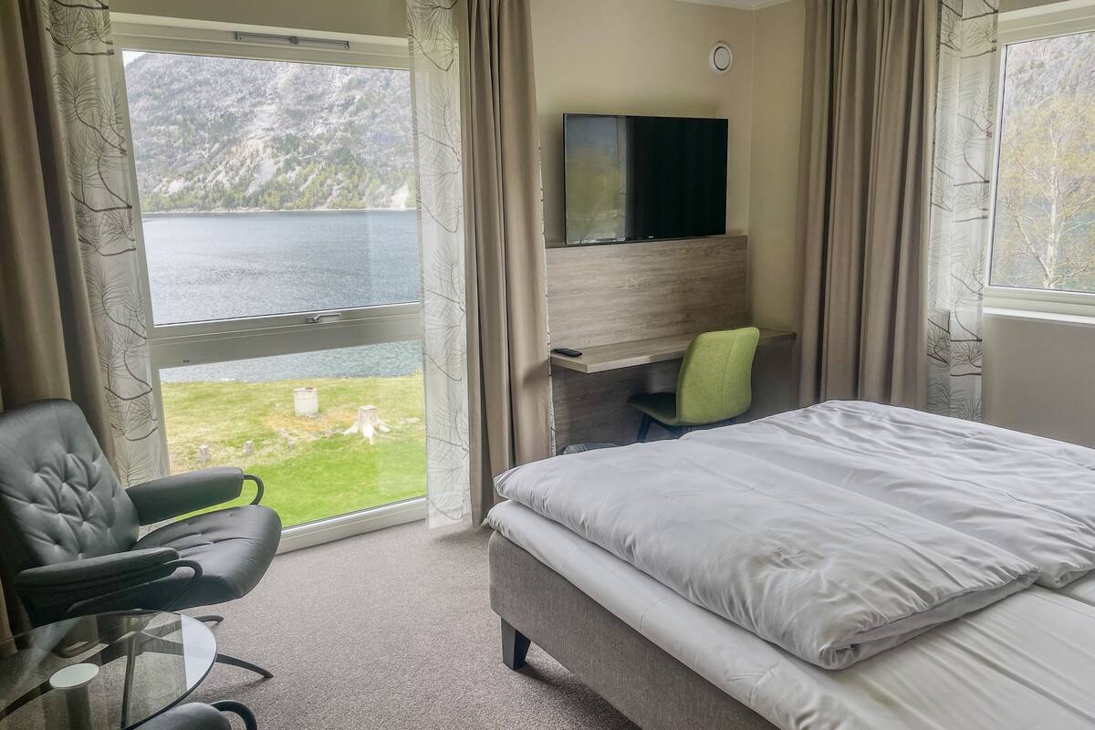 A hotel room with a bed, a desk with a chair, a TV, and a view of a lake.