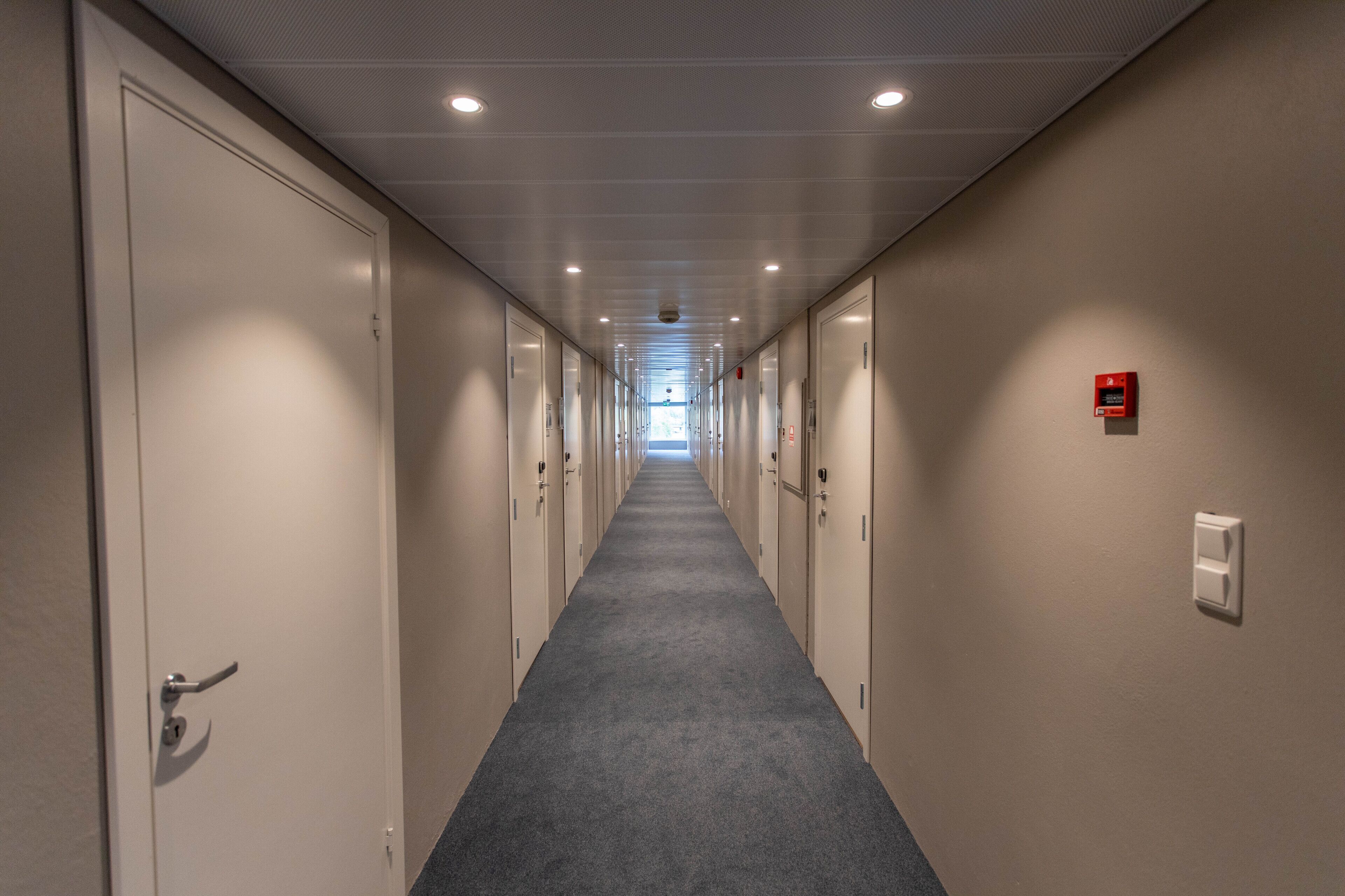 A long, narrow hallway with doors on both sides, leading to a bright room at the end.