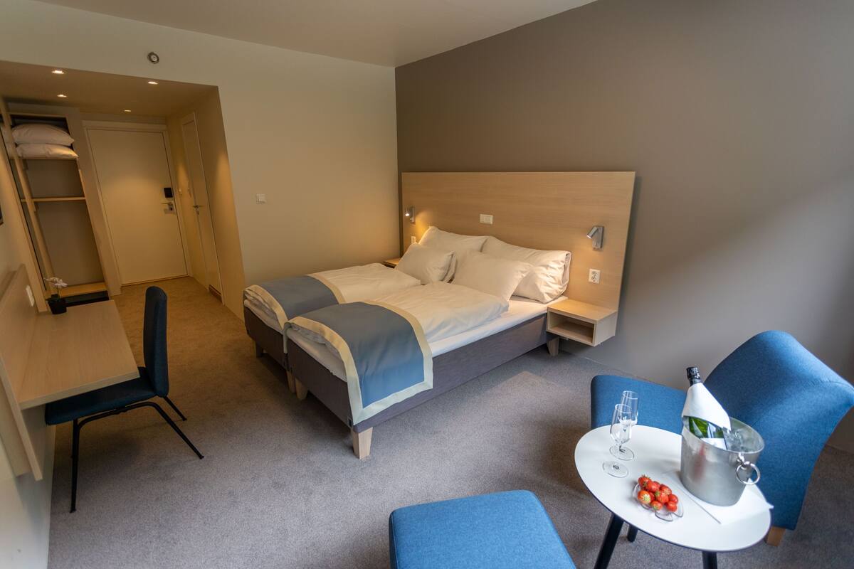 A hotel room with a double bed, a desk with a chair, a small table with a bottle and glasses, and a blue armchair.