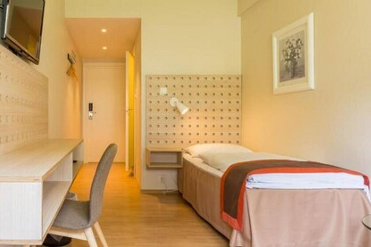A hotel room with a bed, a desk, a chair, a bedside table, and a wall-mounted shelf.