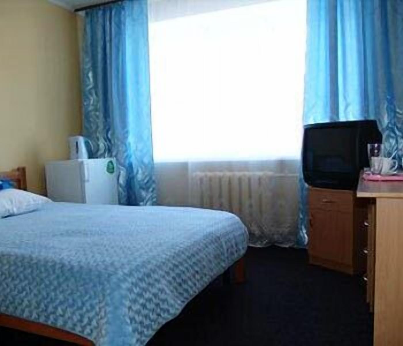 A hotel room with a bed, a television, a small table with a pink object, and a window with blue curtains.