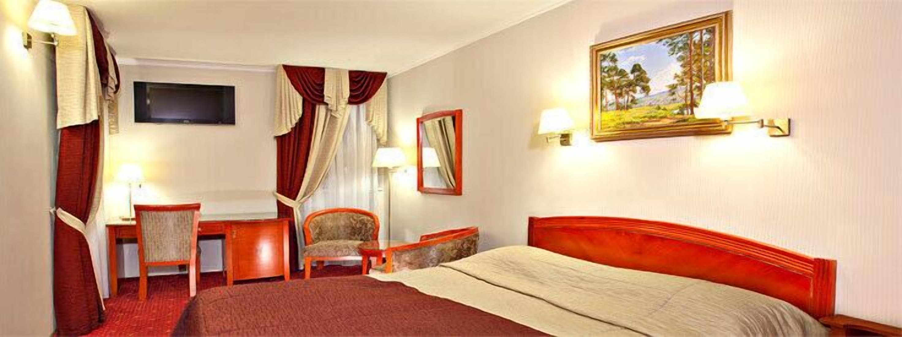 A hotel room with a bed, desk, chair, and a painting on the wall.