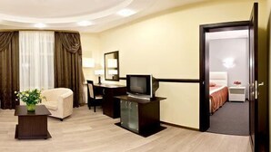 Quarto de hotel com sofá, poltrona, televisão e escrivaninha com cadeira.
