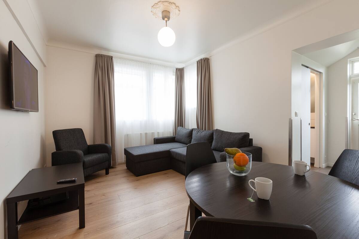 A modern living room with a sectional sofa, a dark wooden coffee table, a TV, and a dining table with a fruit bowl and cups.