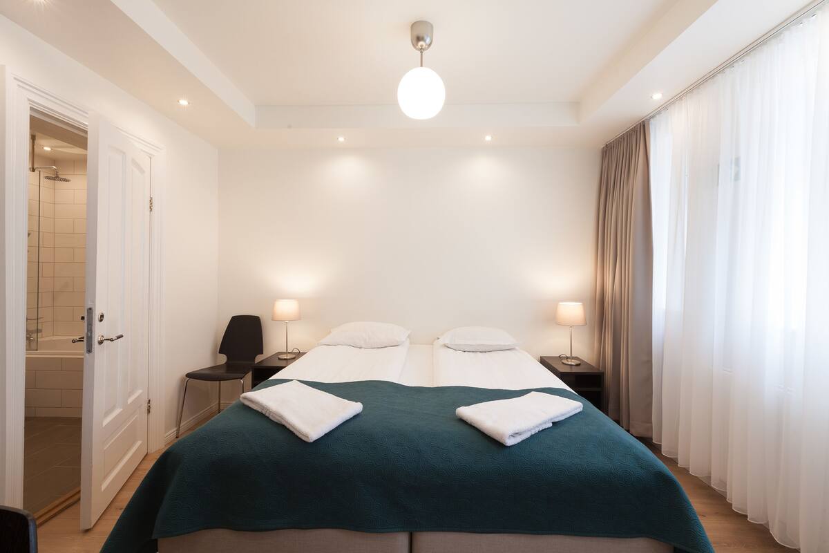 A hotel room with two beds, a shower, a chair, and a nightstand with a lamp.