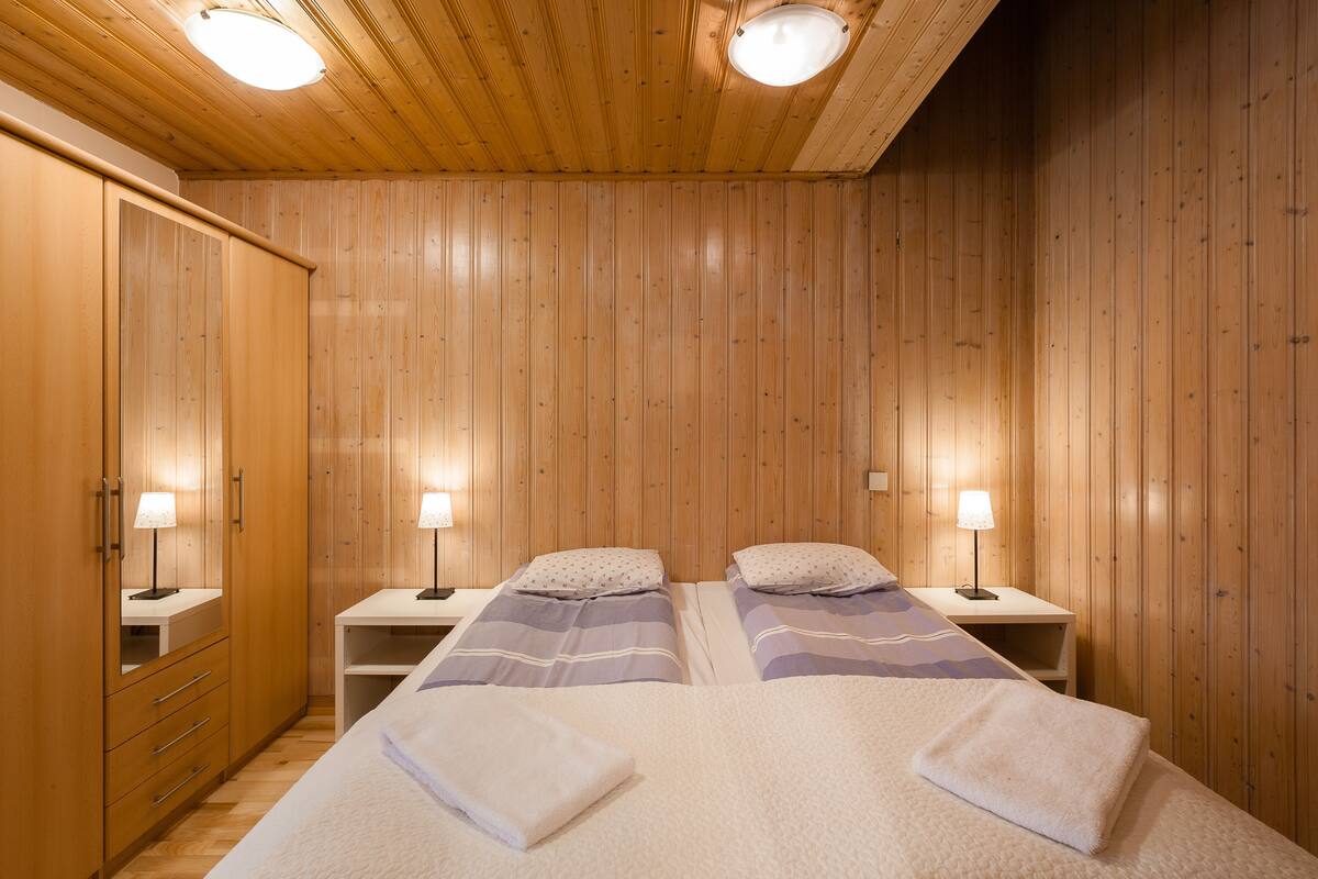 A room with wooden panel walls, two beds with white and purple bedding, nightstands with lamps, and a wardrobe with drawers.