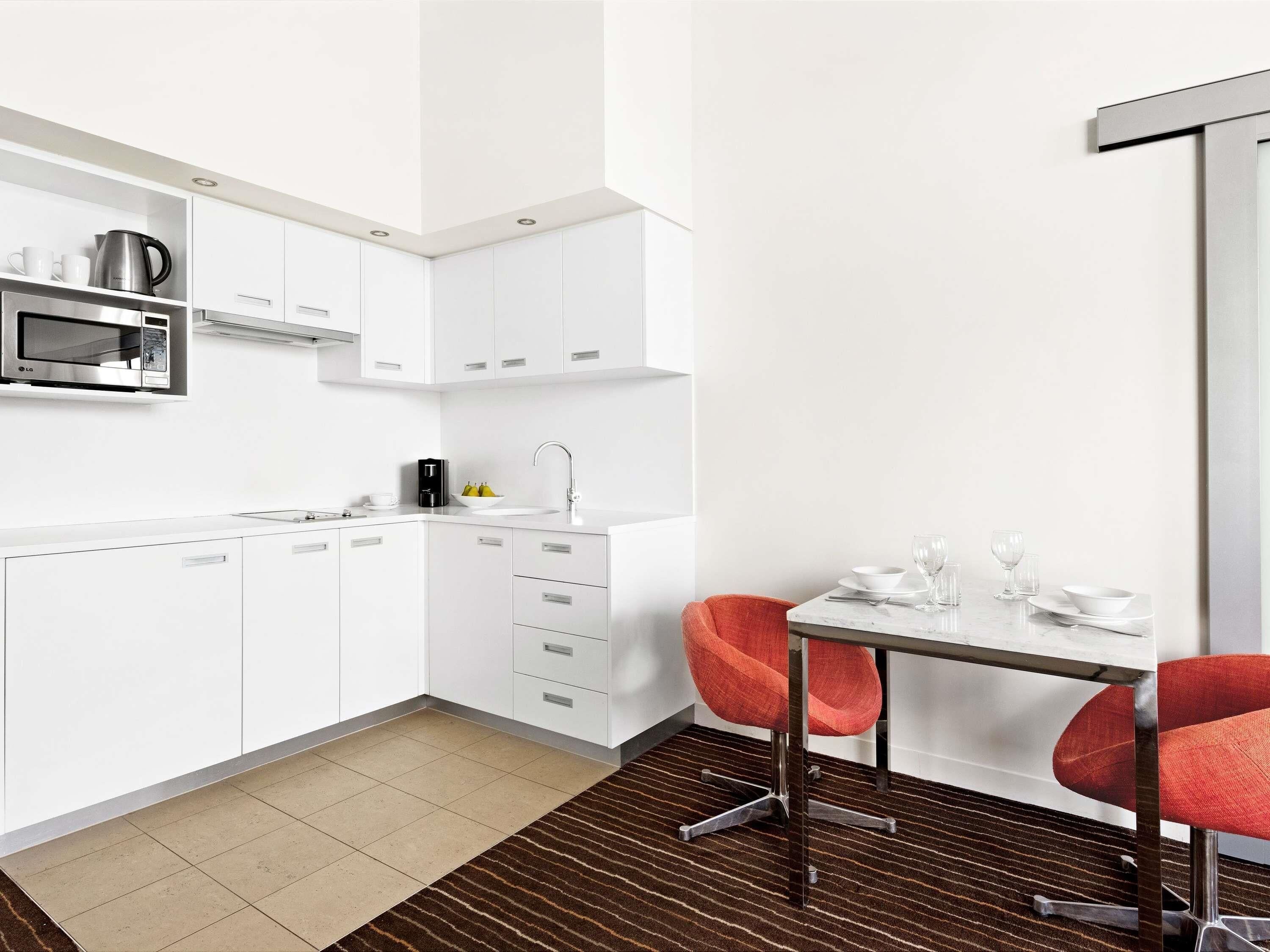 Een moderne keuken met witte kasten, een magnetron en een koffiezetapparaat. Er is een eethoek met een tafel en twee rode stoelen.