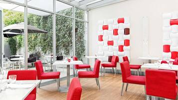 A modern restaurant with red and white chairs, glass tables, and a view of greenery through large windows.