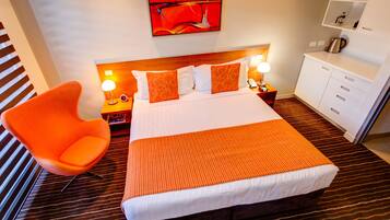 A hotel room with a large bed, an orange chair, a nightstand with a lamp, and a small cabinet.