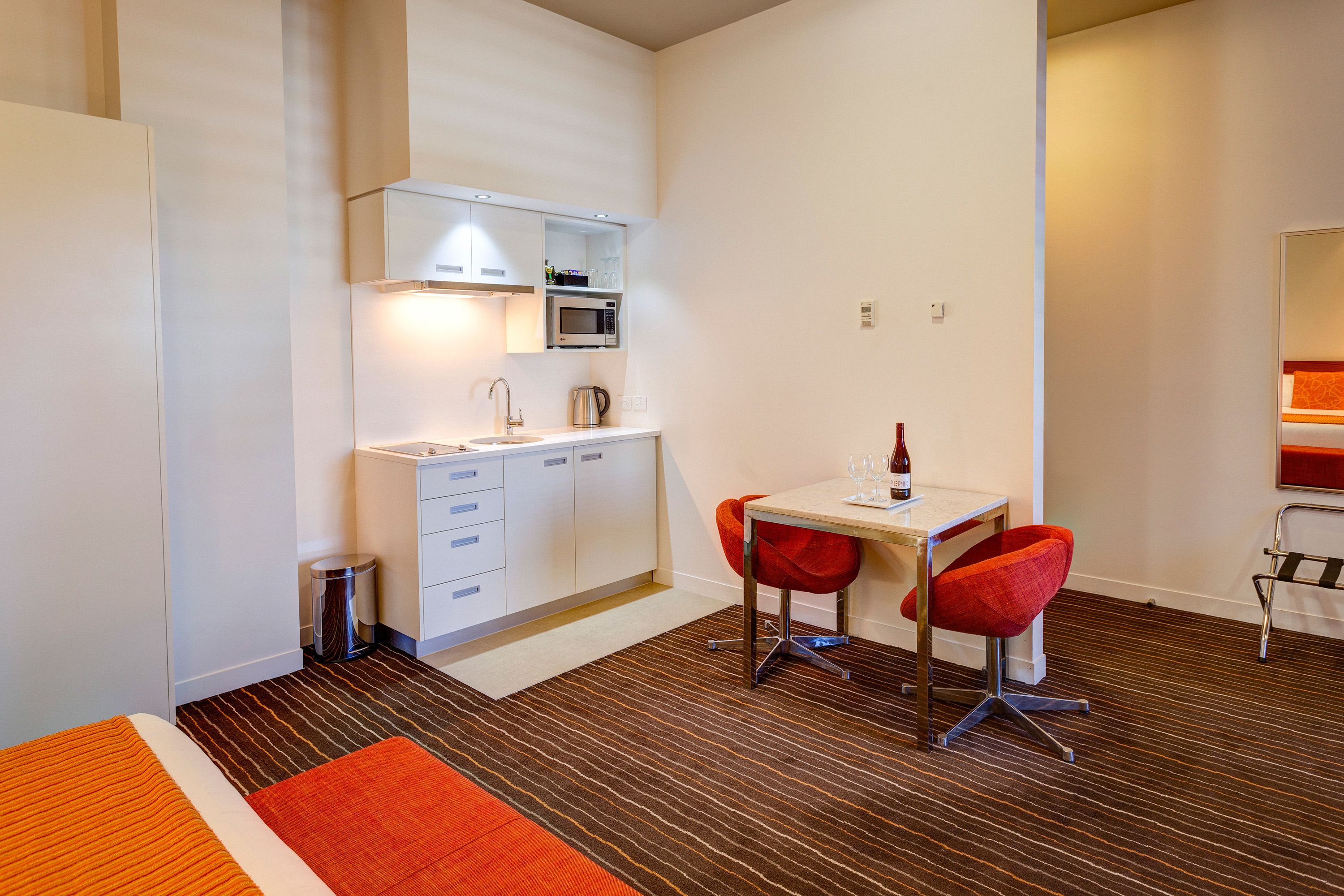Una habitación de hotel compacta con kitchenette, un comedor con dos sillas rojas y un pequeño cubo de basura.