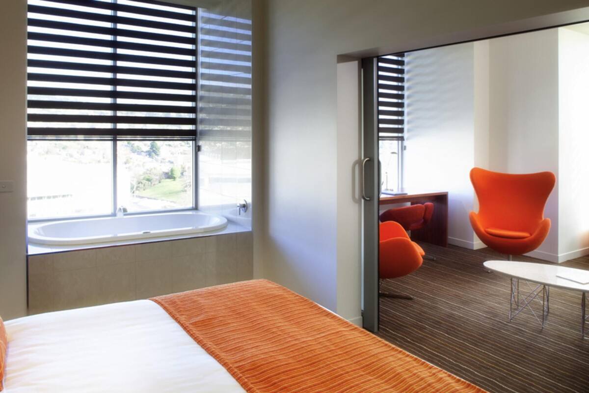 A hotel room with a bed, a window with blinds, a bathroom with a sink and bathtub, and a seating area with a chair and table.