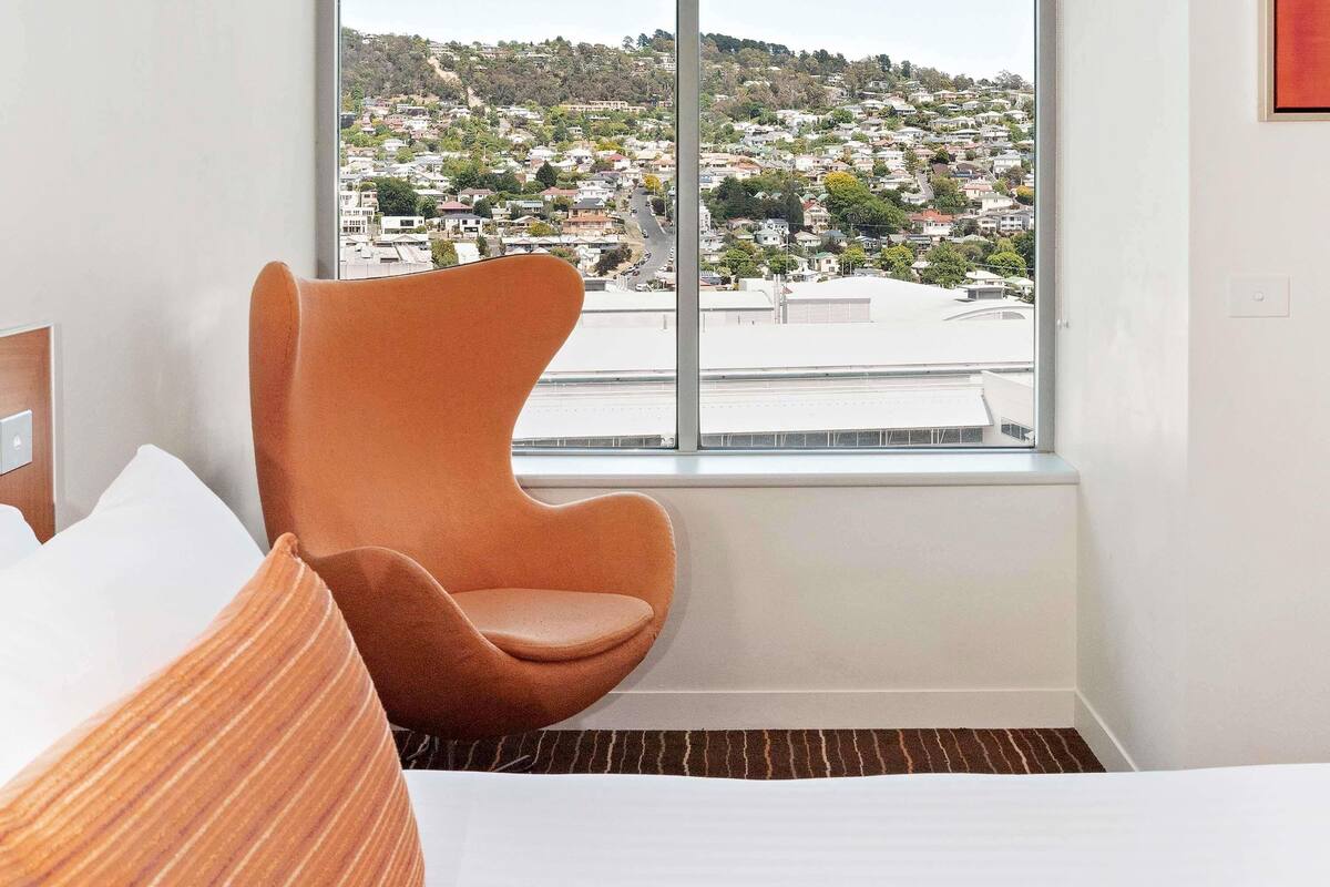 A hotel room with a large window, a bed with white linens, and a unique orange chair.