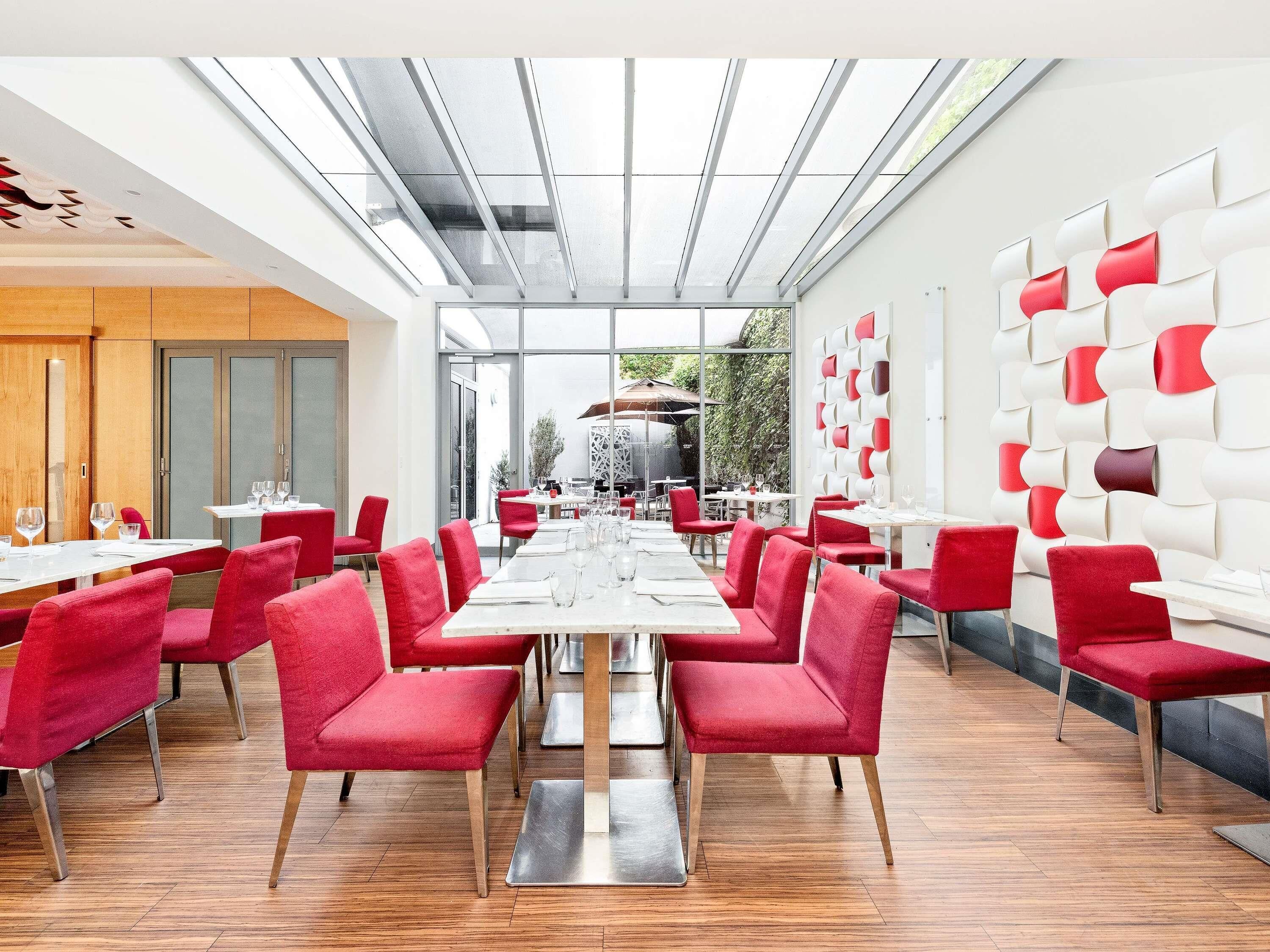 A modern dining area with red chairs and white tables, a glass ceiling, and a view of the outdoors.
