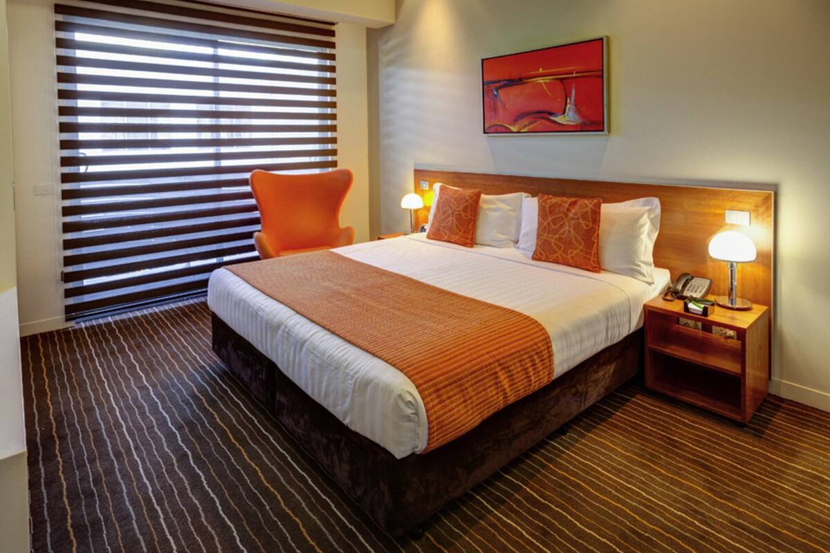 A hotel room with a large bed, a nightstand with a lamp, a chair, a window with blinds, and a painting on the wall.