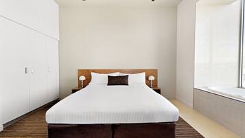 A neatly made bed in a hotel room with a large window, a wardrobe, and a bedside table with lamps.