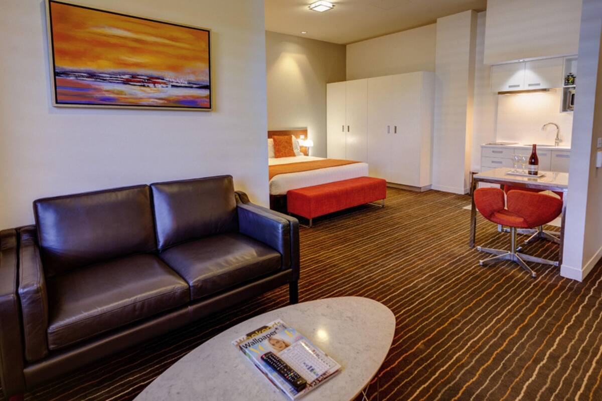 A hotel room with a brown leather sofa, a bed with a red accent, a dining area with a red chair, and a painting on the wall.