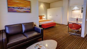 A hotel room with a brown leather sofa, a bed with a red accent, a dining area with a red chair, and a painting on the wall.