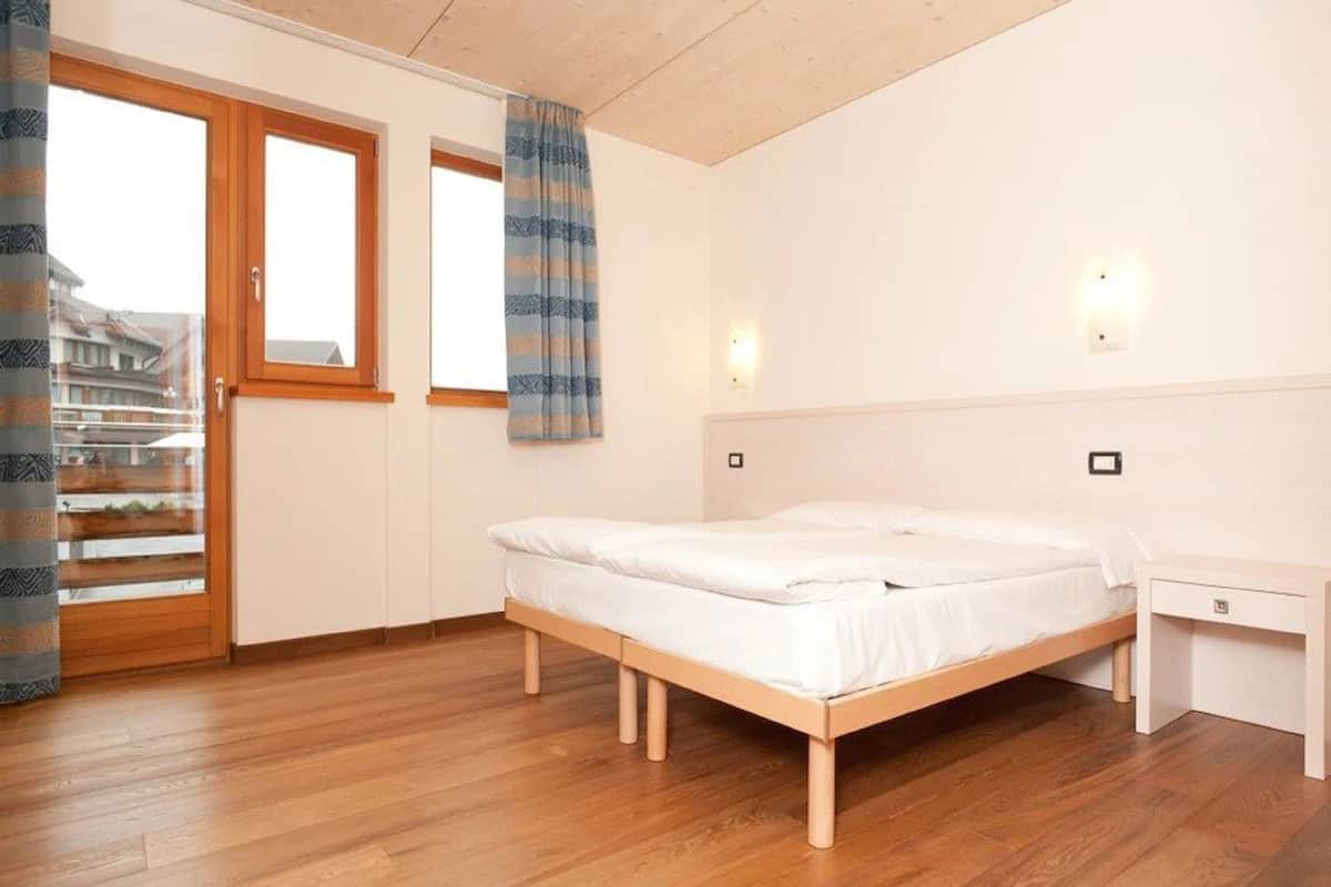 A bedroom with a wooden bed, a nightstand, a window with curtains, and a view of buildings outside.