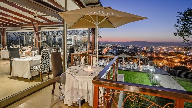 An outdoor dining area with tables and chairs, a large umbrella, and a view of the city at dusk. - Francolin Lodge (Mbombela)