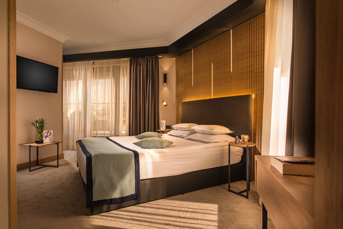 A modern hotel room with a large bed, bedside tables, a TV, and a view of the city.