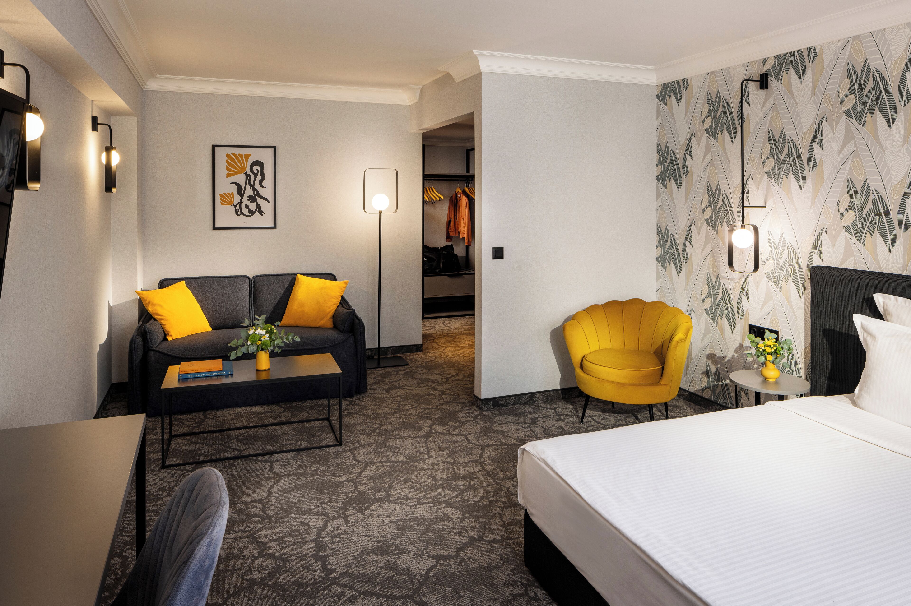 Une chambre d’hôtel avec un lit, un canapé avec des coussins jaunes, une petite table avec un vase, un fauteuil jaune, une armoire et un mur orné d’un motif de feuilles.