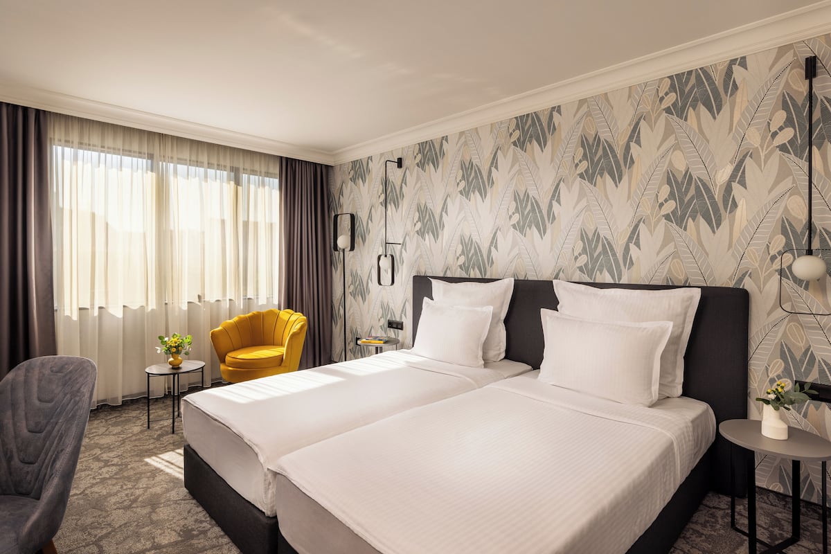 A hotel room with two beds, a yellow armchair, a small table with a vase, and a large window with curtains.