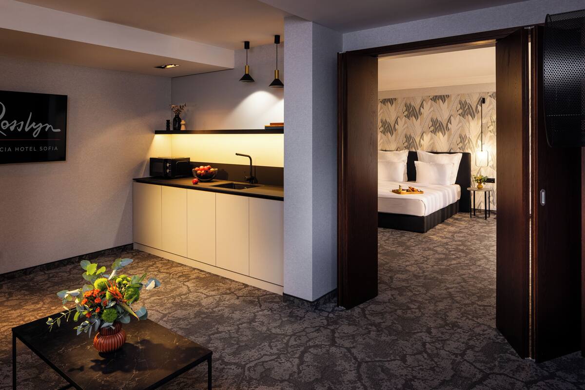 A hotel room with a kitchenette, a bed, a small table with a vase, and a door leading to another room.