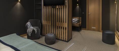 A modern bedroom with a bed, a TV mounted on a wooden panel, a chair, a small round ottoman, and a wardrobe with a mirror.