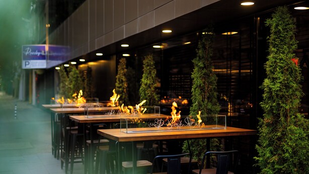 Un espacio exterior para comer con mesas y sillas, rodeado de vegetación e iluminado con luz cálida.