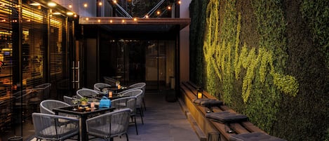 Un espacio exterior para comer con sillas y mesas de mimbre, luces colgantes y un jardín vertical de color verde.