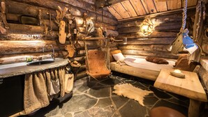 A rustic log cabin interior with a stone floor, a wooden bench, a bed, and a small table with a candle. - Engholm Husky Design Lodge (Karasjok)