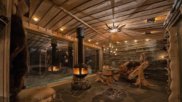 A cozy interior with a stone floor, wooden beams, a central fireplace, and a large window. - Engholm Husky Design Lodge (Karasjok)