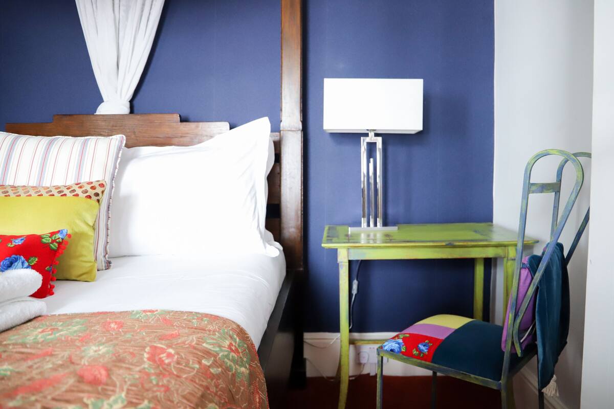 A neatly made bed with a canopy, a bedside table with a lamp, a chair with a colorful cushion, and a wall with a blue accent.
