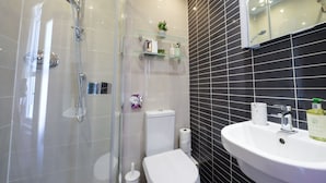 A modern bathroom with a glass-enclosed shower, a white toilet, a sink with a mirror above it, and a black tiled wall. - The Cavalaire (Brighton)