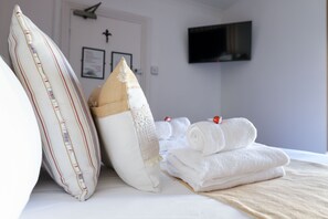 A neatly made bed with pillows and folded towels, a flat-screen TV mounted on the wall, and a framed certificate or award on the door. - The Cavalaire (Brighton)