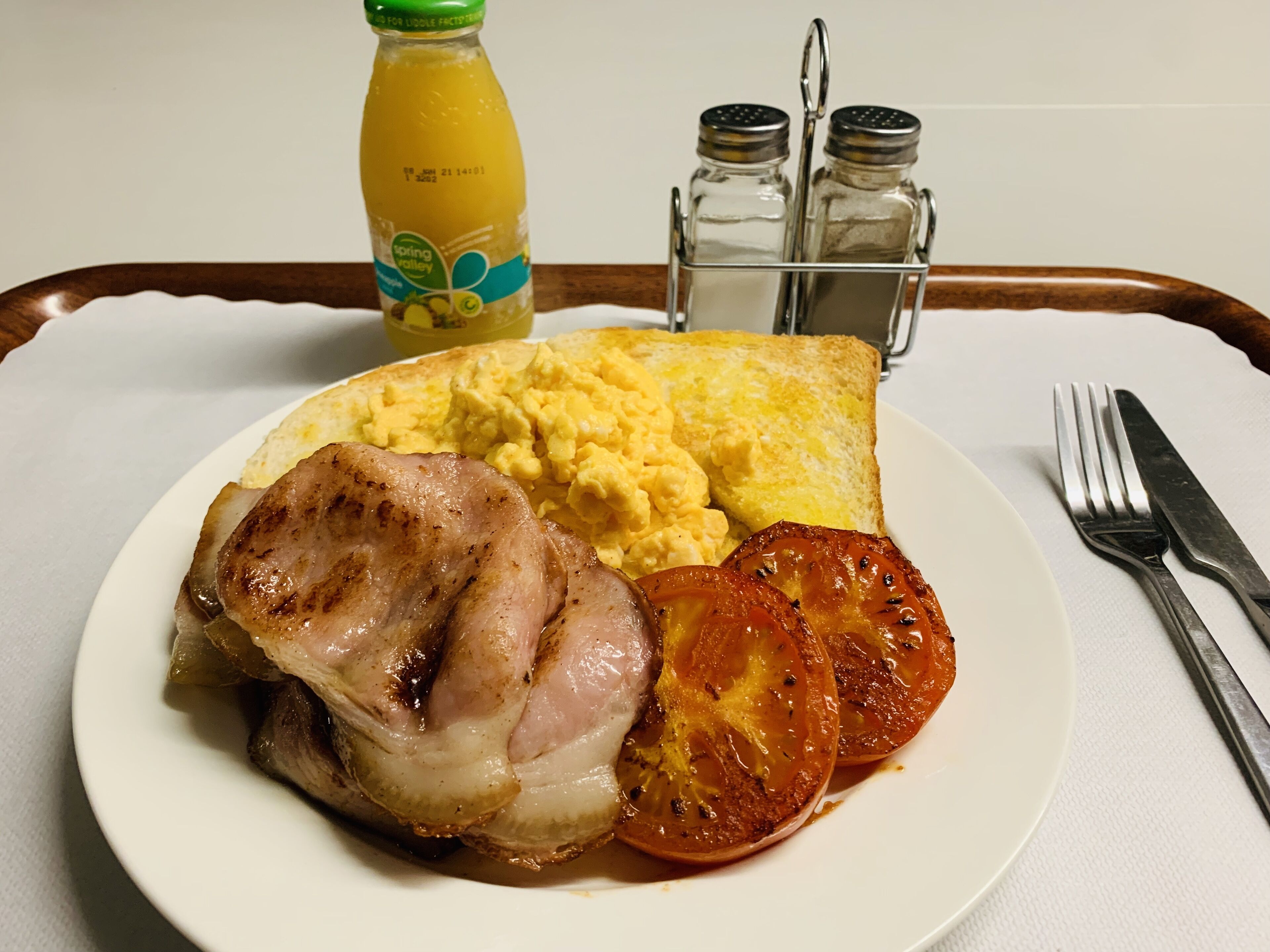A plate with grilled pork, scrambled eggs, and tomato slices, accompanied by a bottle of orange juice and salt and pepper shakers on a tray.