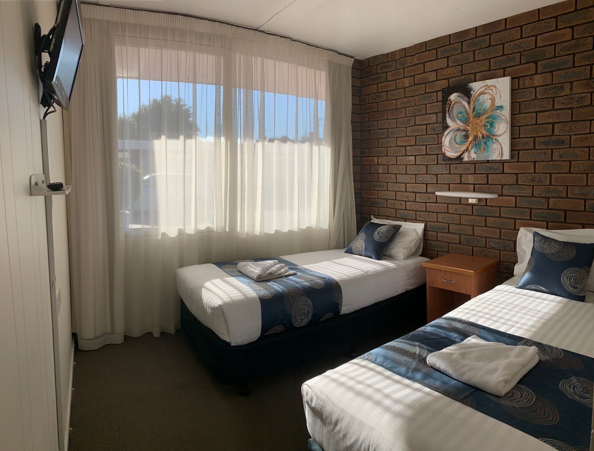A hotel room with two beds, a TV mounted on the wall, a brick accent wall, and a window with sheer curtains.