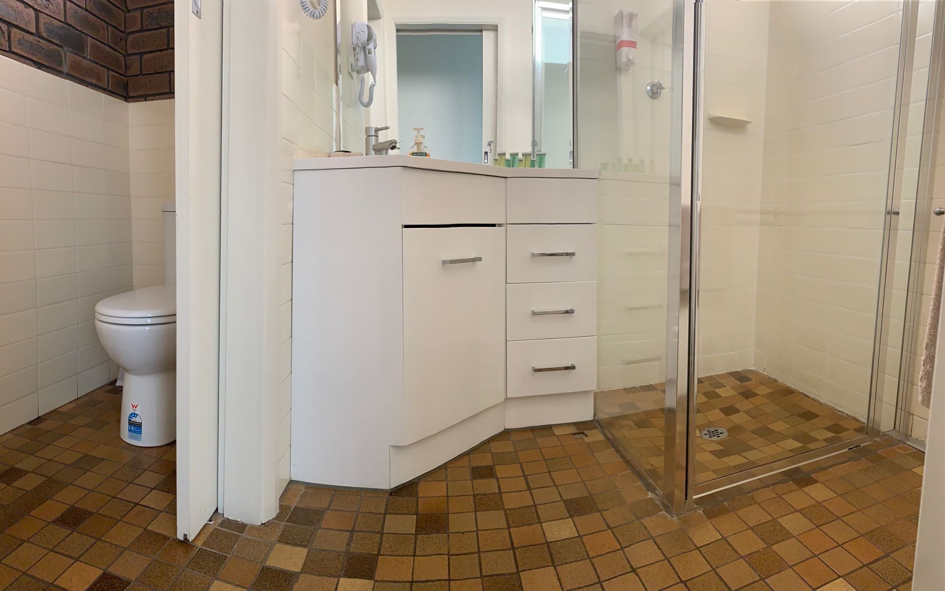 A bathroom with a toilet, a white vanity with drawers, a glass shower enclosure, and a tiled floor.