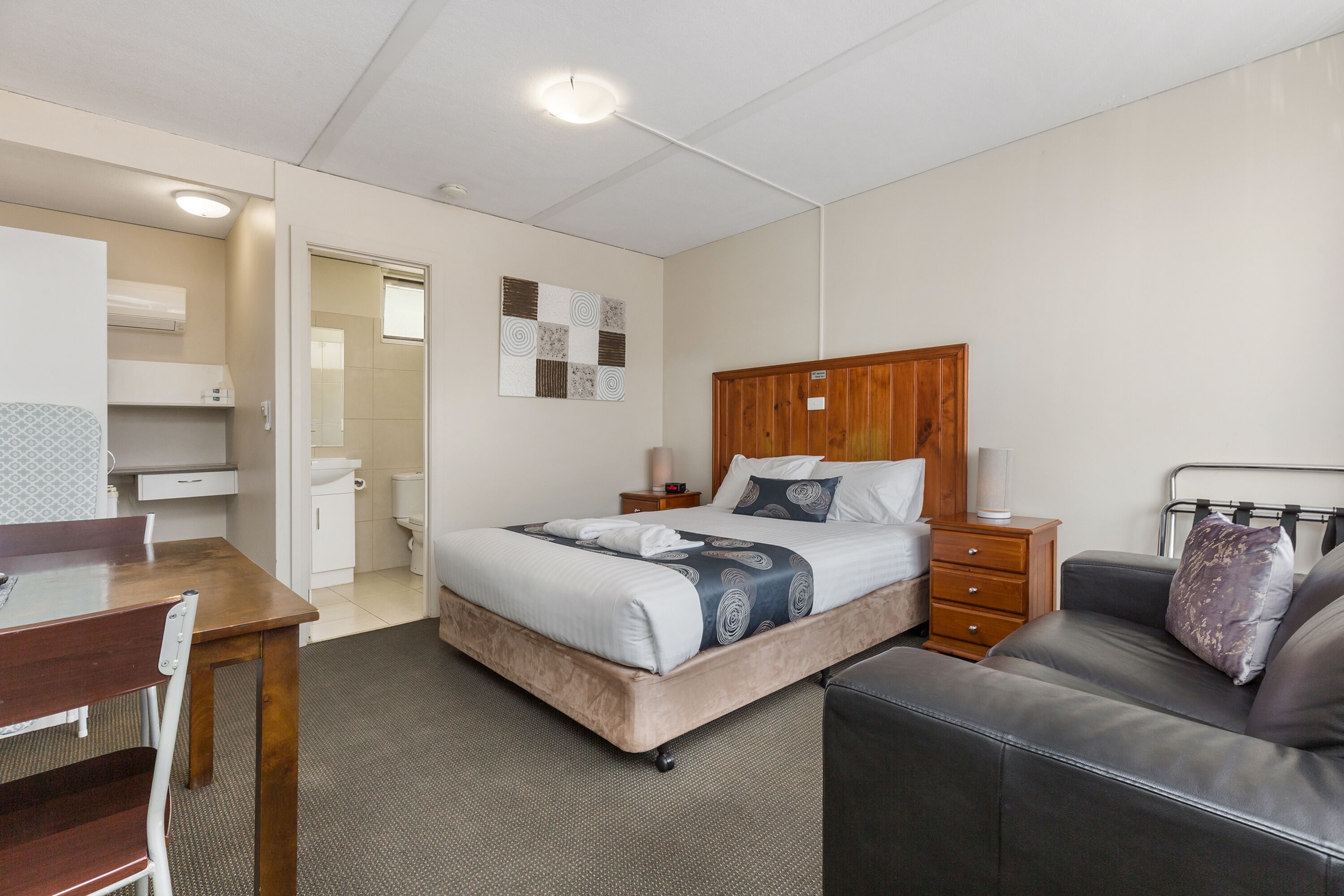 A hotel room with a bed, a desk, a chair, a sofa, a wooden headboard, and a wall-mounted artwork.