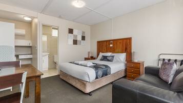 A hotel room with a bed, a desk, a chair, a sofa, a wooden headboard, and a wall-mounted artwork.