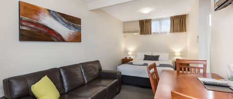 A hotel room with a leather sofa, a wooden dining table, a bed with a white duvet, and a painting on the wall.