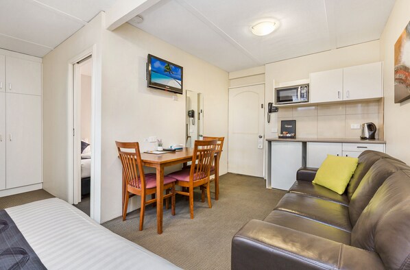 A compact living space with a kitchenette, a dining area with wooden chairs, a sofa, and a TV mounted on the wall. - Bendigo McIvor Motor Inn (East Bendigo)