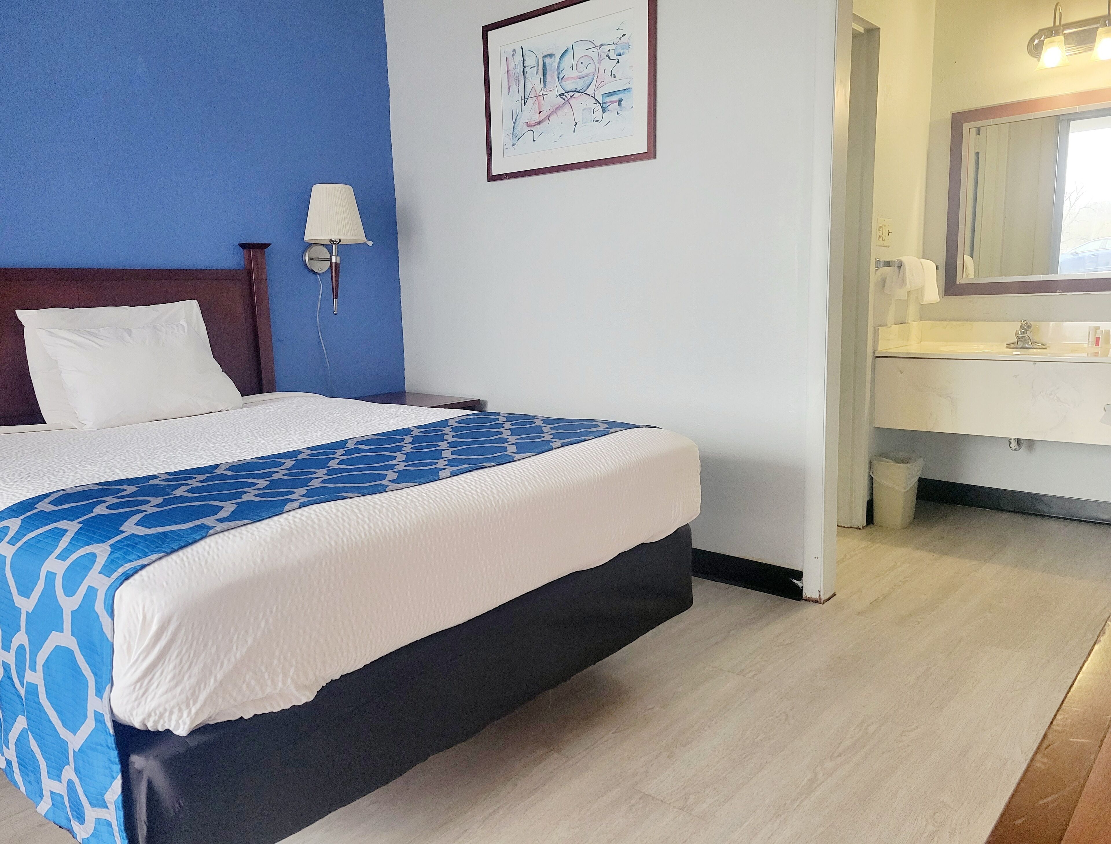 A hotel room with a bed, a nightstand, a wall-mounted lamp, a framed artwork, and a bathroom with a sink and mirror.