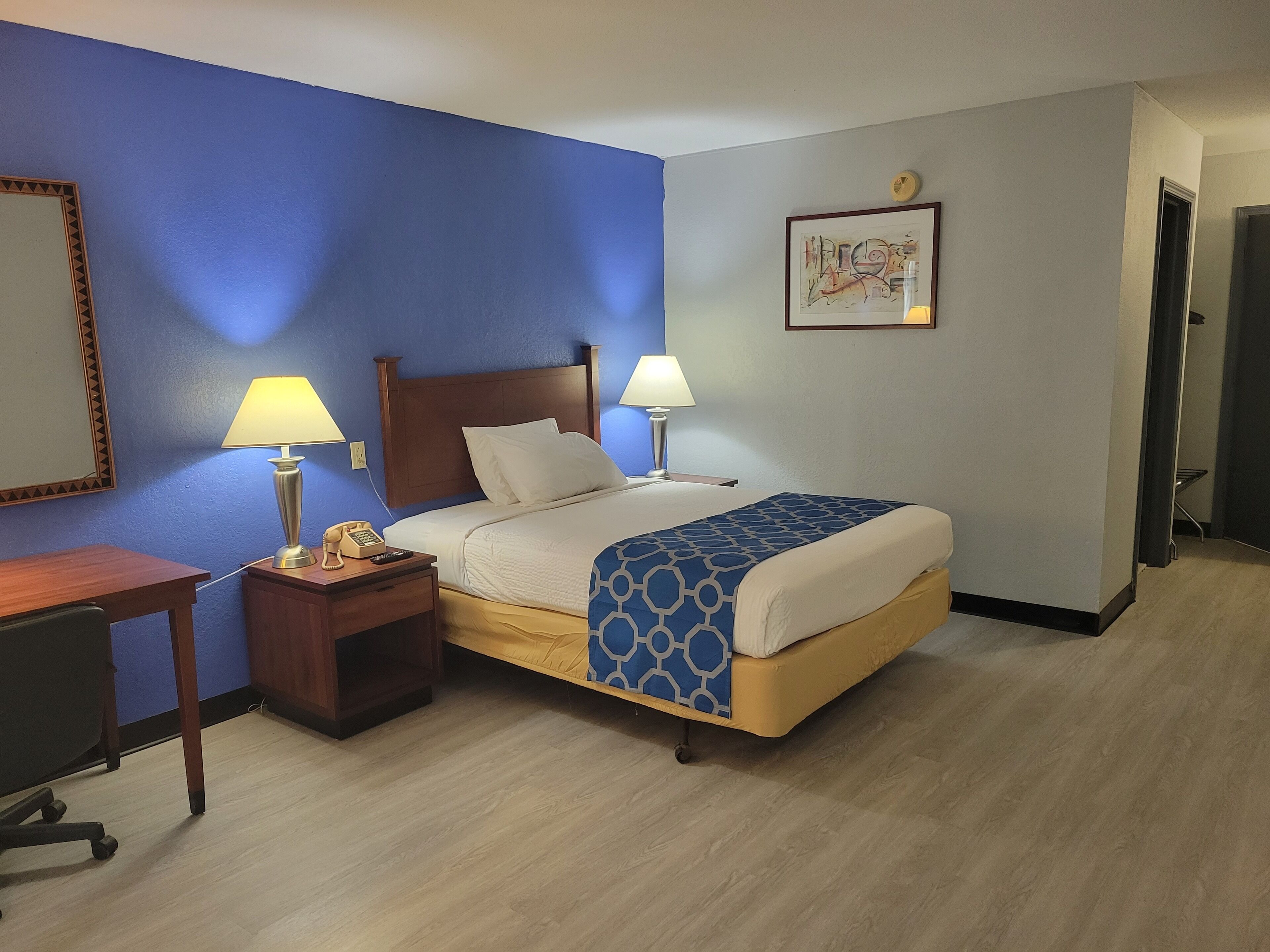 A hotel room with a bed, two bedside lamps, a desk with a chair, a telephone, and a framed picture on the wall.