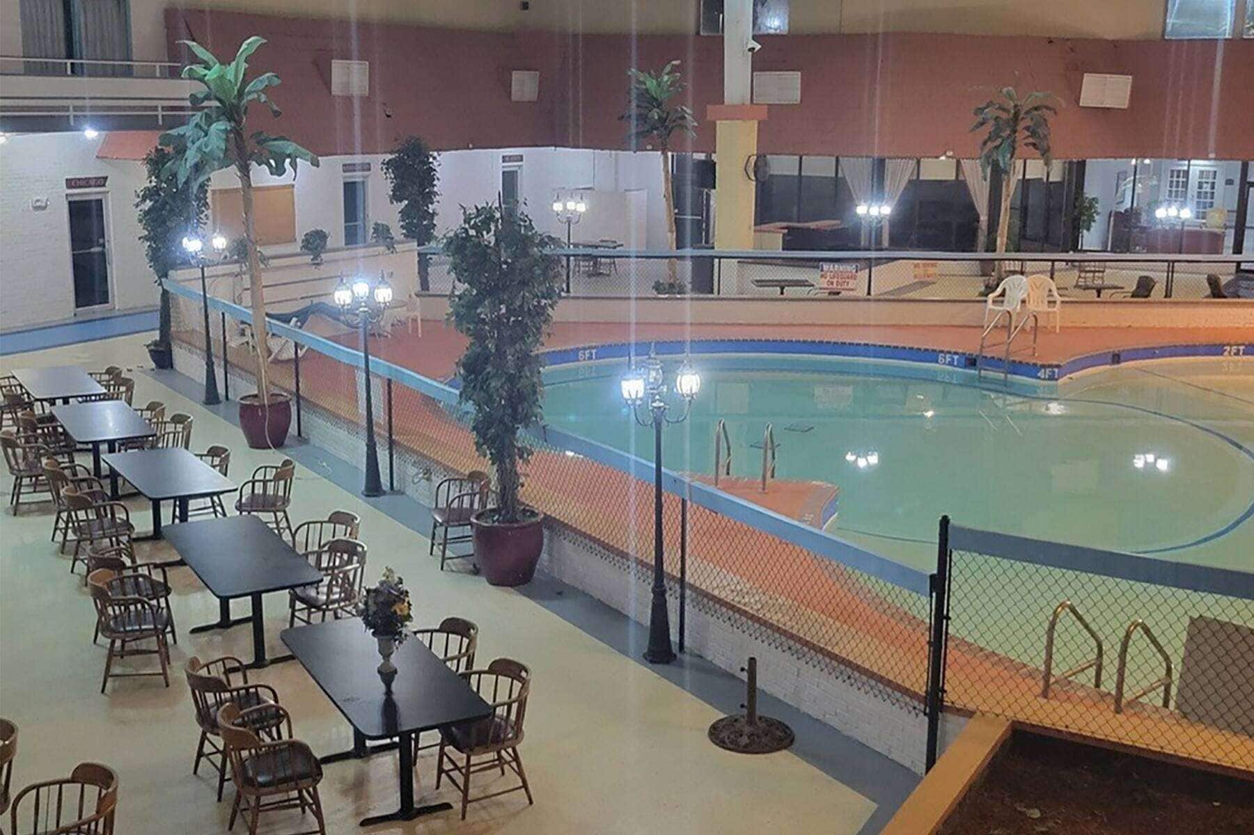 An indoor swimming pool area with tables and chairs, surrounded by a fence, and a basketball court visible in the background.