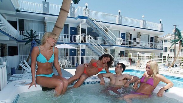 Ett poolområde med en bubbelpool, solstolar och en byggnad med balkonger i bakgrunden.